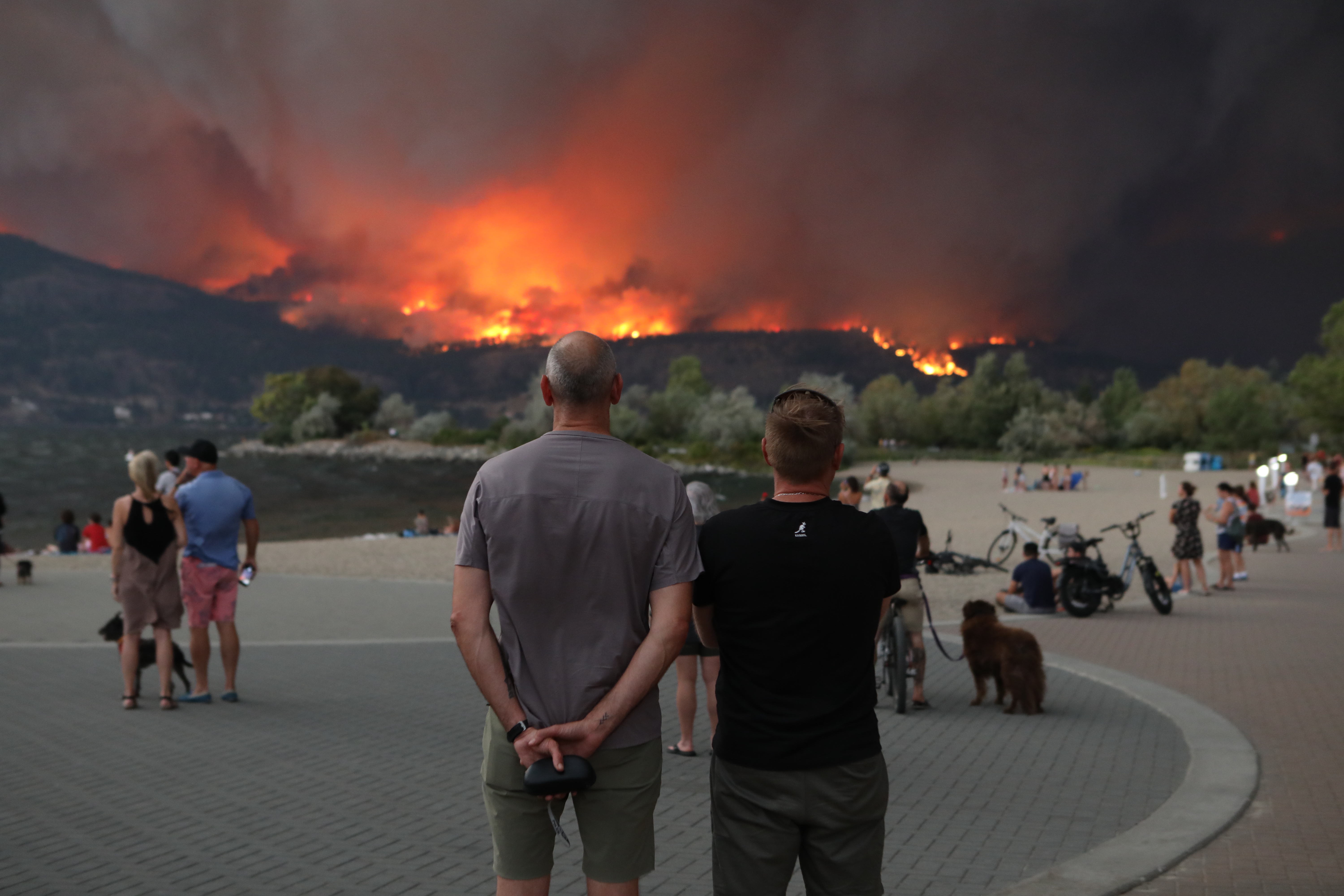 People watch from downtown Kelowna, B.C., as the McDougall Creek wildfire burns in the hills above West Kelowna, B.C., on the evening of Aug. 17, 2023. The blaze, which measured at 1,100 hectares that same night, has resulted in thousands of evacuation alerts, and led to the the City of West Kelowna and parts of Westbank First Nation declaring a state of emergency that earlier that day. (Photo by Aaron Hemens for the Globe and Mail)