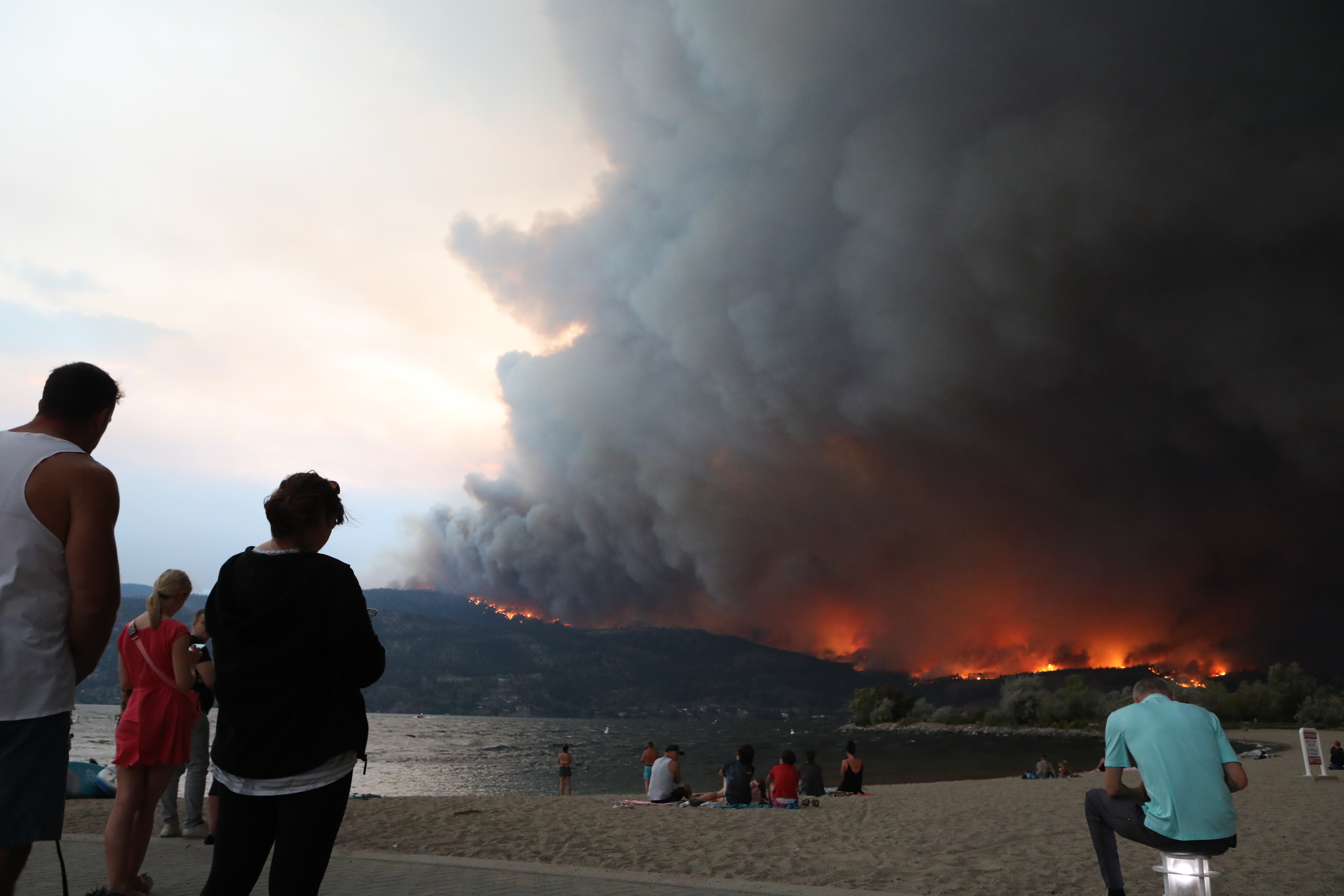 People watch from downtown Kelowna, B.C., as the McDougall Creek wildfire burns in the hills above West Kelowna, B.C., on the evening of Aug. 17, 2023. The blaze, which measured at 1,100 hectares that same night, has resulted in thousands of evacuation alerts, and led to the the City of West Kelowna and parts of Westbank First Nation declaring a state of emergency that earlier that day. (Photo by Aaron Hemens for the Globe and Mail)