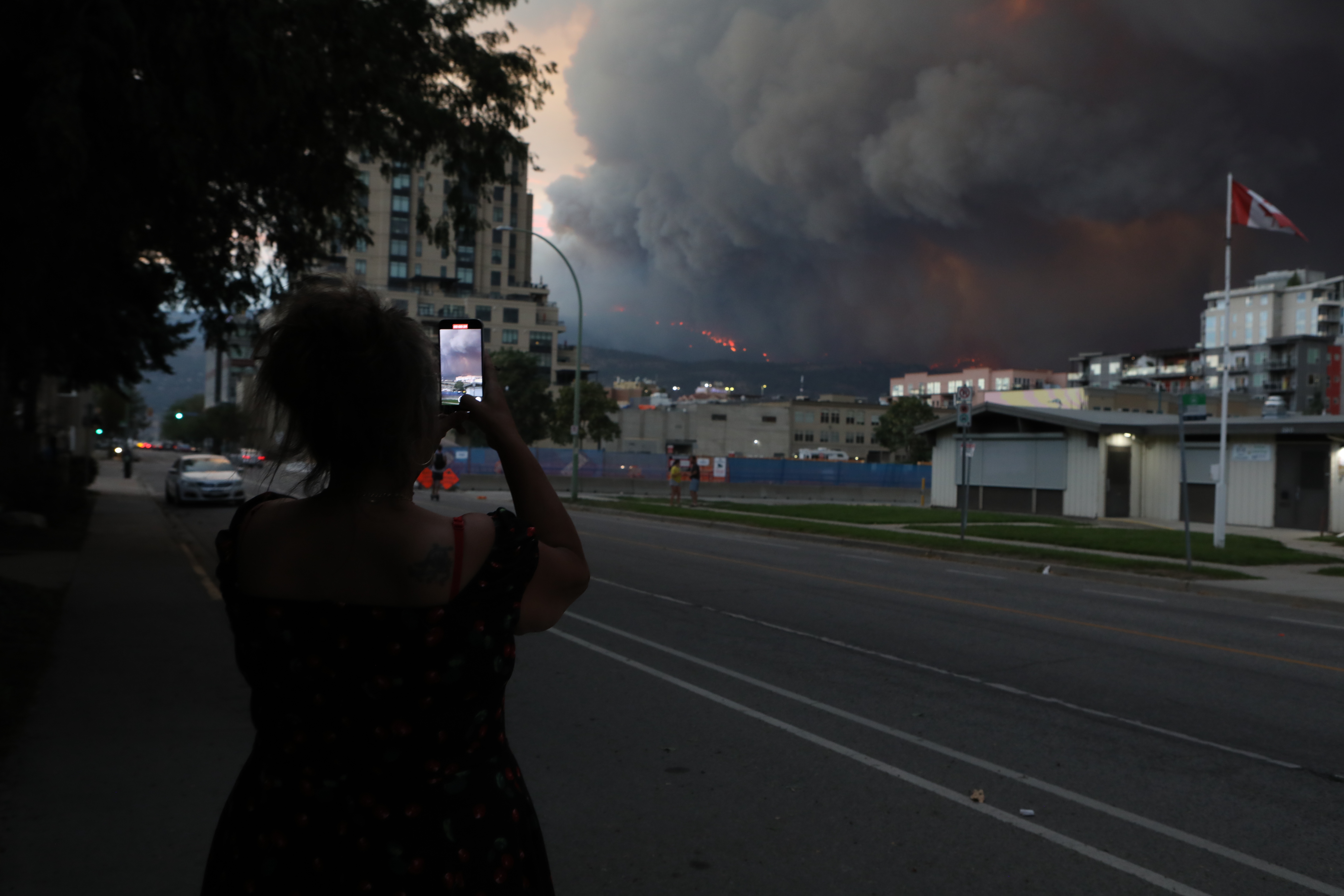 A person takes a photo from downtown Kelowna, B.C., of the McDougall Creek wildfire burning in the hills above West Kelowna, B.C., on the evening of Aug. 17, 2023. The blaze, which measured at 1,100 hectares that same night, has resulted in thousands of evacuation alerts, and led to the the City of West Kelowna and parts of Westbank First Nation declaring a state of emergency that earlier that day. (Photo by Aaron Hemens for the Globe and Mail)